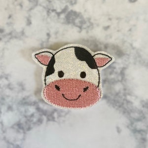 May include: A white and black cow face with pink nose and mouth, embroidered with glitter on a white fabric.