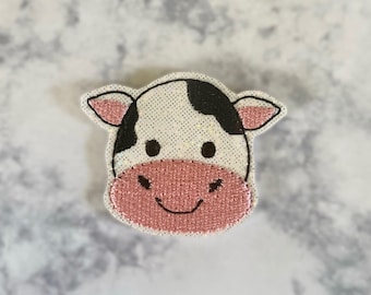 May include: A white and black cow face with pink nose and mouth, embroidered with glitter on a white fabric.