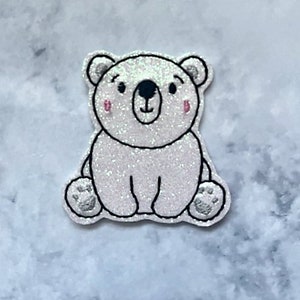 May include: A white glitter polar bear applique with black eyes and a black nose. The bear is sitting with its paws in front of it.