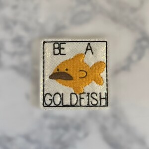 Może przedstawiać: Haftowana naszywka z kreskówkową złotą rybką i brązowym wąsem. Kwadratowa naszywka zawiera czarny napis "BE A GOLDFISH". Ryba jest pomarańczowa, na białym, błyszczącym tle.