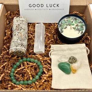 May include: A gift box with the words "Good Luck Harmony + Positivity + Abundance" on the lid. The box contains a sage smudge stick, a clear crystal, a green beaded bracelet, a small bag with green, gold, and clear crystals, and a black candle with green and gold crystals embedded in the wax.