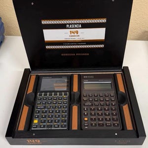Könnte beinhalten: Eine schwarze Holzkiste mit Klappdeckel, die zwei Taschenrechner enthält. Die Box trägt ein Etikett mit der Aufschrift "PLASENCIA 149 COSECHA PRIVADA". Die Taschenrechner sind dunkelgrau mit zahlreichen Tasten und Bildschirmen. Der Innenraum hat holzfarbene Akzente.