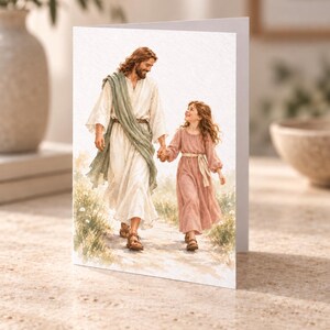 May include: A greeting card featuring a watercolour illustration of a long-haired man in a white robe with a sage green sash walking hand-in-hand with a young girl in a dusty rose dress through a field of small white flowers on a dirt path.