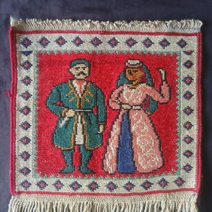May include: A square woven textile wall hanging featuring a red centre panel with two figures in traditional attire. The left figure wears a teal coat, and the right figure wears a pink patterned dress. The border has a diamond pattern with fringe.