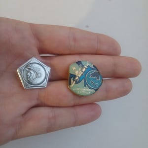 Puede incluir: Dos pines de metal. Uno es un pin pentagonal de tono plateado que presenta el perfil de un cosmonauta con casco y el texto CCCP. El otro es un pin circular con esmalte azul y verde que representa un cohete y un globo terráqueo con el texto СОТРУДНИЧЕСТВО В КОСМОСЕ.