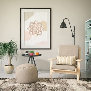 Op de afbeelding: Een ingelijste wandkunstprint met een bruin mandala-ontwerp op een cr&egrave;mekleurige achtergrond met abstracte vormen. De print hangt boven een licht houten fauteuil met een gestreept kussen. Een kleine bijzettafel met boeken, een plant en een mok staat in de buurt.