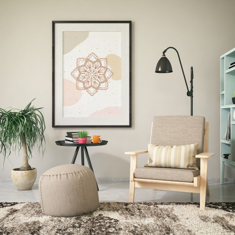 Op de afbeelding: Een ingelijste wandkunstprint met een bruin mandala-ontwerp op een cr&egrave;mekleurige achtergrond met abstracte vormen. De print hangt boven een licht houten fauteuil met een gestreept kussen. Een kleine bijzettafel met boeken, een plant en een mok staat in de buurt.