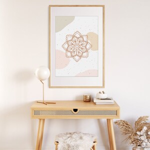 Op de afbeelding: Een ingelijste print met een bruin mandala-ontwerp op een witte achtergrond met beige en roze abstracte vormen. De print hangt boven een houten bureau met een kleine lamp, een kaars en een decoratieve vaas. Een kruk met een zacht wit kussen staat eronder.