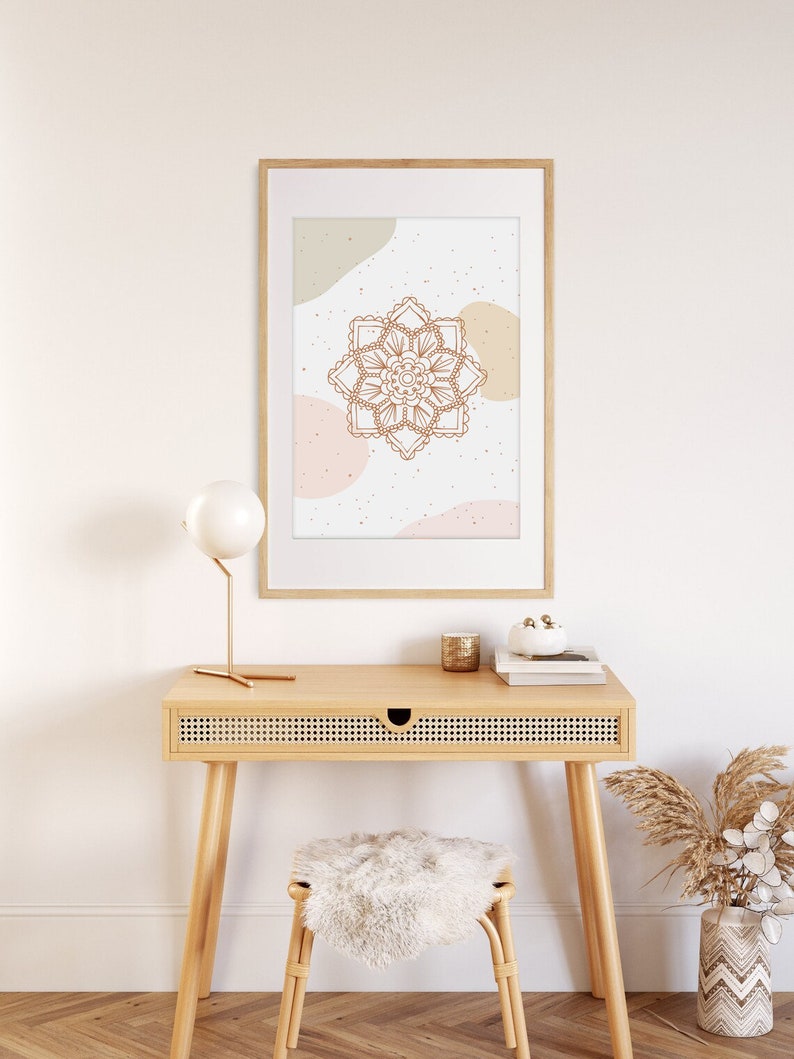 Op de afbeelding: Een ingelijste print met een bruin mandala-ontwerp op een witte achtergrond met beige en roze abstracte vormen. De print hangt boven een houten bureau met een kleine lamp, een kaars en een decoratieve vaas. Een kruk met een zacht wit kussen staat eronder.