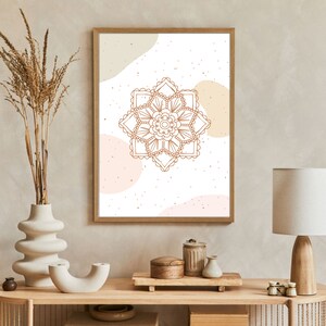 Op de afbeelding: Een ingelijste print met een bruin mandala-ontwerp op een witte achtergrond, met beige en roze abstracte vormen. Het kunstwerk wordt tentoongesteld op een houten console met decoratieve items, waaronder een vaas met gedroogde planten en een tafellamp.
