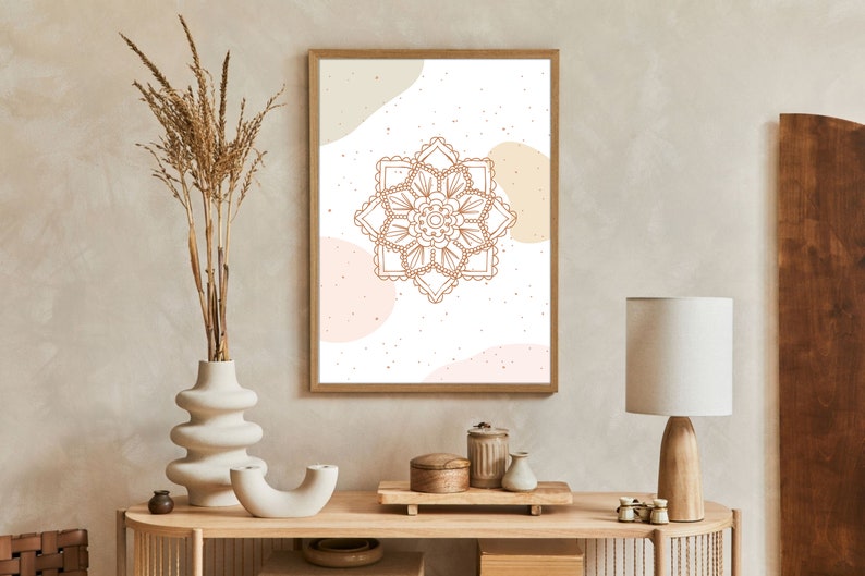 Op de afbeelding: Een ingelijste print met een bruin mandala-ontwerp op een witte achtergrond, met beige en roze abstracte vormen. Het kunstwerk wordt tentoongesteld op een houten console met decoratieve items, waaronder een vaas met gedroogde planten en een tafellamp.