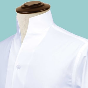 Può includere: Primo piano di una camicia bianca su un manichino. La camicia presenta un colletto alla coreana e una chiusura con un solo bottone. Il tessuto appare liscio e la camicia è ben rifinita, suggerendo uno stile formale o professionale.