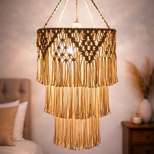 May include: A three-tiered macrame chandelier with a warm, neutral color palette. The light fixture features intricate knotting and fringe details, creating a bohemian aesthetic. The image also shows a bedroom setting with a bed and nightstand.
