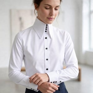 May include: A crisp white long-sleeve button-down shirt featuring a high, structured collar and a front placket adorned with small, round black buttons. The cuffs also feature matching black buttons, creating a sharp contrast against the fabric.