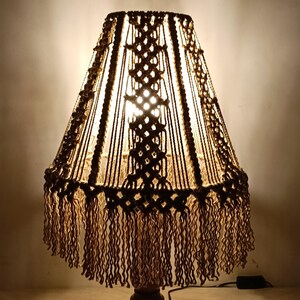 Op de afbeelding: Een handgemaakte macramé lamp met een bruine houten voet en een gefranjerde lampenkap. De kap heeft ingewikkelde knoop patronen in bruin en beige, wat een warme gloed creëert. De lamp is een decoratieve verlichtingsarmatuur.