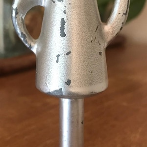 May include: A small, silver-colored metal teapot figurine with a handle and spout. The teapot sits atop a pedestal base. The paint is chipped, revealing dark spots. The figurine is likely a decorative item.