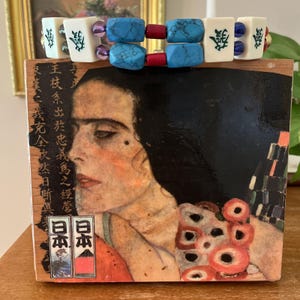 Puede incluir: Un bloque de madera con un retrato de una mujer con cabello largo y negro y un vestido rojo y naranja. El bloque está coronado con una pulsera hecha de cuentas turquesas, rojas y blancas, y fichas de mahjong. Texto japonés visible.