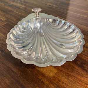 May include: A polished silver-toned metal serving dish shaped like a scallop shell. The dish features a scalloped edge and a small, raised circular knob at the top center. The surface is reflective with deep, radiating grooves that mimic the natural texture of a seashell, creating a bright, metallic appearance.
