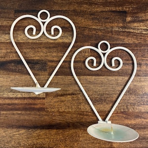 May include: Two white metal wall-mounted candle holders, each featuring a heart-shaped frame with decorative scrollwork at the top and a circular base plate designed to hold a single candle. The frames are constructed from thin, curved metal rods.