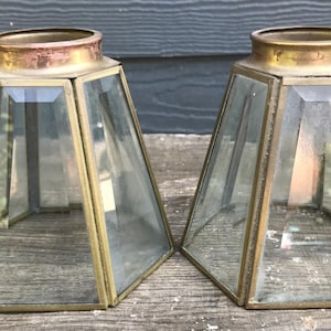 May include: Two vintage glass and brass lamp shades. Each shade has a trapezoidal shape with clear glass panels framed by brass. The top of each shade has a copper-colored ring. The shades are displayed on a wooden surface.