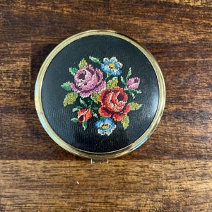 May include: A round compact mirror with a gold-toned metal frame and a black fabric cover featuring a central needlepoint floral design. The embroidery displays pink, red, and blue flowers with green leaves, centered on the circular lid.