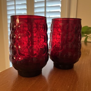 May include: Two ruby red drinking glasses with a textured, bubble-like pattern. The glasses have a short, dark base and a cylindrical shape. The glasses are made of glass and are suitable for beverages.