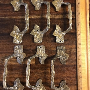 Op de afbeelding: Zeven messingkleurige kastgrepen met bladvormige bases en getextureerde, wijnstokachtige handgrepen. De grepen zijn gerangschikt op een houten oppervlak. Een liniaal is zichtbaar.