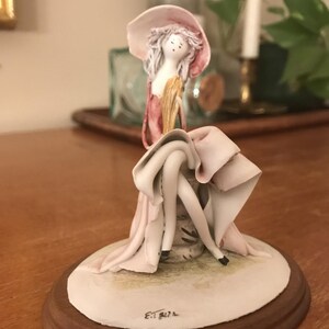 May include: A porcelain figurine of a woman in a pink dress and hat, seated on a white base with a wooden rim. The figurine has delicate features and is painted with soft colors. The woman's legs are crossed, and her dress is draped around her.