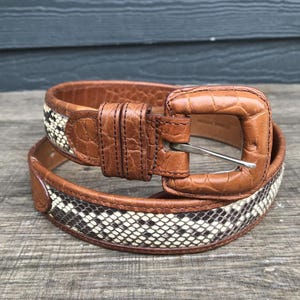 May include: A brown leather belt with a snakeskin pattern. The belt has a square buckle and a silver-colored metal pin. The snakeskin pattern is black and white. The belt is on a wooden surface.