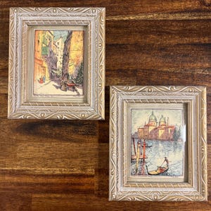 May include: Two rectangular wall frames with ornate, light-colored, carved borders. The left frame contains a print of a street scene with figures, while the right frame displays a print of a Venetian canal with a gondola and domed buildings.