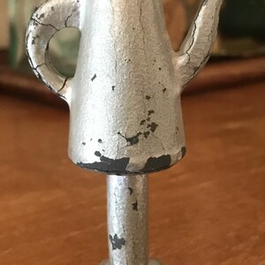 May include: A small, silver-colored metal teapot figurine with a pedestal base. The teapot has a handle and spout, and the paint is chipped, revealing a darker color underneath. The figurine stands approximately 4 inches tall.