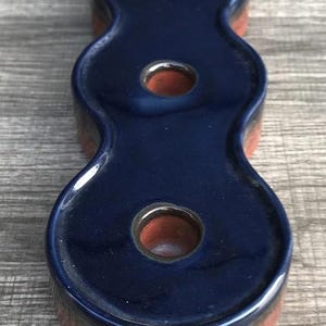 May include: A navy blue ceramic candle holder with three holes. The candle holder has a unique, elongated shape with a glossy finish. The base and the inside of the holes are a reddish-brown color.