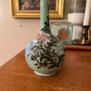May include: A light green ceramic vase with a long, slender neck and a rounded body. The surface features a detailed hand-painted design of dark brown branches and foliage, accented with small, textured pink and blue floral elements.