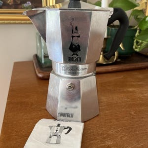 Puede incluir: Una cafetera italiana Bialetti Moka Express de color plateado con asa y parte superior negras. La imagen icónica de un hombre con bigote está en la parte delantera. Un pequeño folleto de instrucciones está al lado.