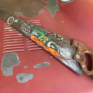 May include: A hand saw with a wooden handle and a blade decorated with a racing car theme. The blade features a green and orange design with the number 13, a black cat, and flames.