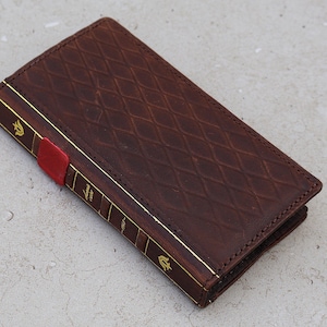 Può includere: Un portafoglio in pelle marrone con motivo a rombi e finiture dorate, simile a un libro. Il portafoglio ha un segnalibro rosso ed è chiuso. Il design è classico ed elegante.