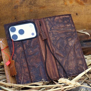 May include: Open brown leather phone case with a textured surface. The case holds a white phone with three dark blue camera lenses. A red cord and a small leather tag are attached. The case is open, revealing the phone and interior.