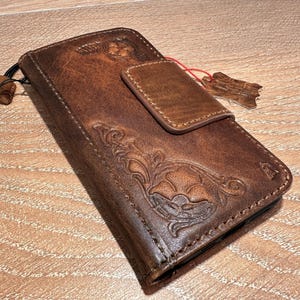 A brown leather wallet or fanny pack with embossed designs and a small red tag attached to it, resting on a wooden surface.