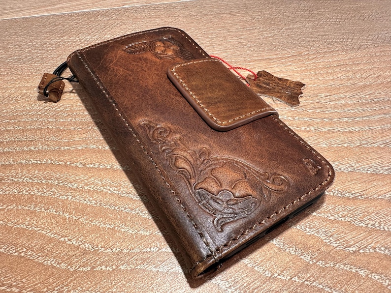 A brown leather wallet or fanny pack with embossed designs and a small red tag attached to it, resting on a wooden surface.