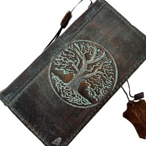 Könnte beinhalten: Dunkelbraune Ledergeldbörse mit geprägtem Lebensbaum-Design und türkisfarbener Patina. Die Geldbörse hat eine rechteckige Form und ein abgenutztes Aussehen. Ein Lederanhänger hängt an der Geldbörse. Das Baumdesign befindet sich in einem Kreis.