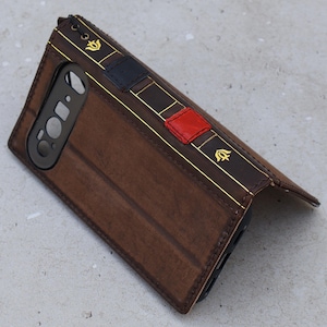 Puede incluir: Una funda de teléfono de cuero marrón con un diseño de libro. La funda presenta detalles dorados, un acento rojo y azul marino y un emblema decorativo. La funda está diseñada para abrirse y tiene un recorte para la cámara.