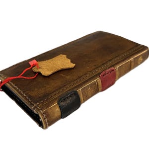 Può includere: Un portafoglio in pelle marrone con un design a libro vintage. Il portafoglio ha una finitura invecchiata, una piccola etichetta in pelle e dettagli rossi, neri e rossi. Il portafoglio è chiuso e sembra nuovo.