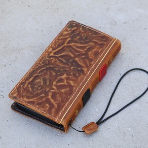 Könnte beinhalten: Eine braune Leder-Handyhülle mit einem strukturierten Blumendesign. Die Hülle hat eine goldene Zierleiste, einen roten Akzent und eine schwarze Handschlaufe. Die Hülle ist geschlossen und hat weiße Nähte.
