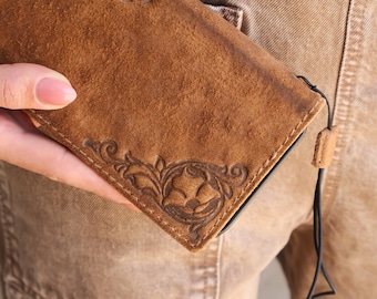Könnte beinhalten: Eine braune Leder-Handyhülle mit einem geprägten Blumendesign in der Ecke. Die Hülle hat ein strukturiertes, Wildleder-ähnliches Aussehen und ein schwarzes Schlüsselband. Die Hülle wird in der Hand einer Person gehalten.