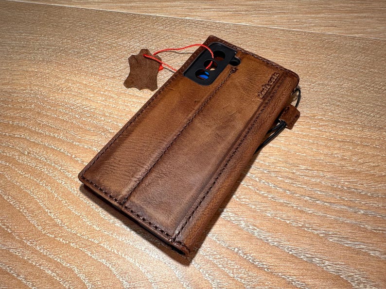 A brown leather wallet or case with a zipper closure and a small red tag attached to it, resting on a wooden surface.