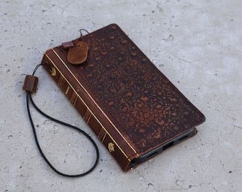 May include: A brown leather phone case with a book-like design. The case features embossed floral patterns, gold trim, and a matching leather tag. A black cord with a wooden clasp is attached for carrying.