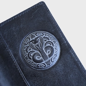 May include: A dark charcoal gray leather wallet featuring a circular embossed design on the front. The embossed emblem depicts a detailed cobra snake surrounded by intricate, symmetrical scrollwork patterns, all highlighted in a lighter gray tone.