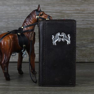 May include: A brown leather wallet with a silver horse design. The wallet has a flap closure and a strap.