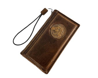 May include: A brown leather wallet with a gold-colored horse emblem. The wallet has a black wrist strap and is rectangular in shape. The leather has a distressed appearance.