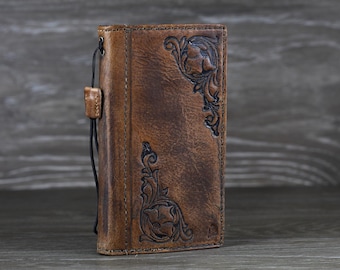 May include: A brown leather wallet with embossed floral designs on the front. The wallet has a distressed finish and a black cord attached to the side. The leather is a warm, medium brown color.